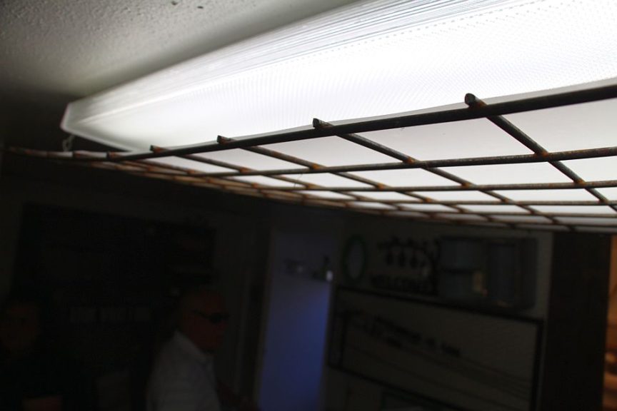 A metal grid is suspended directly below a florescent light, with a thin, frosted sheet of plastic atop it